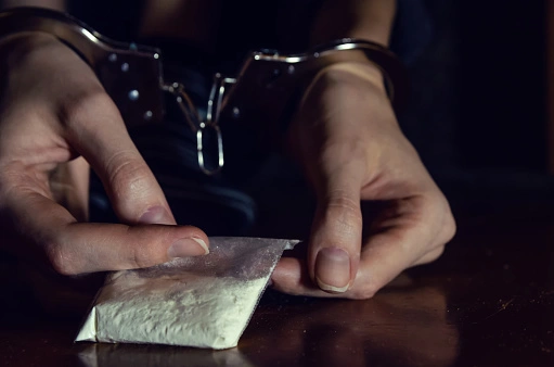 Close up of hands in handcuffs holding a bag of white powder drugs.