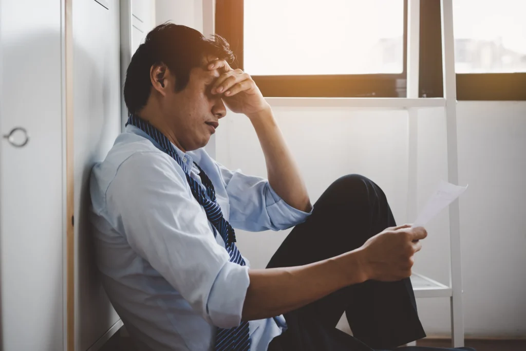 A man holding his head with his tie loosened reading a piece of paper sitting on the floor against the wall.